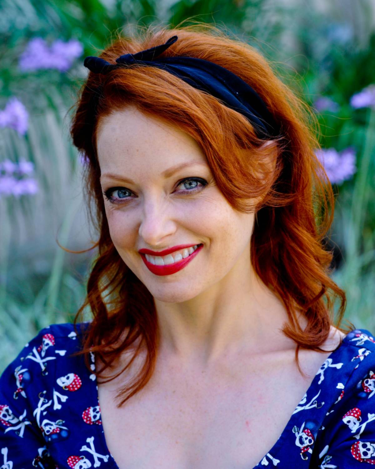 Elizabeth is a caucasian woman with red hair and blue eyes. She is wearing a blue top and is smiling widely at the camera.