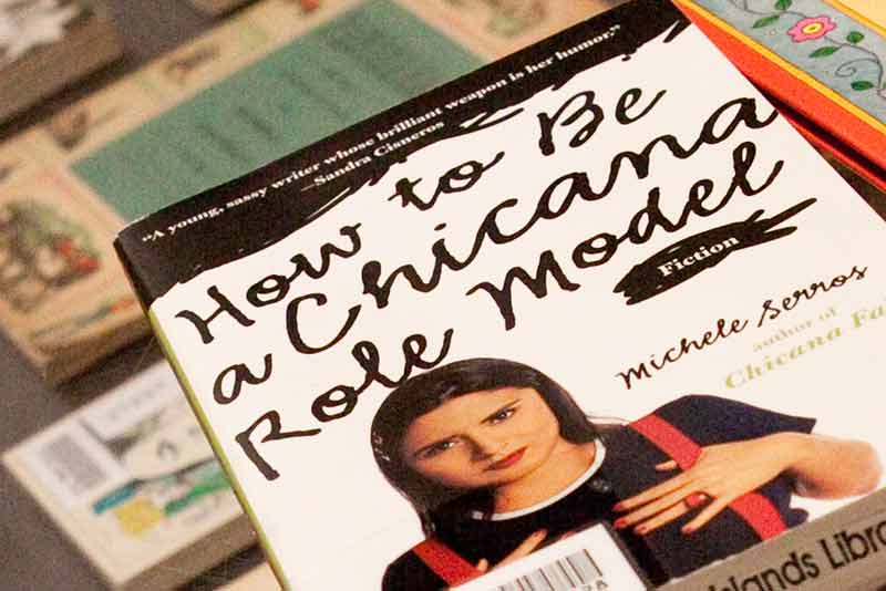 a pile of books with the book How to Be a Chicana Model on top which has a white cover and a Chicana on the cover in a black tshirt and red backpack