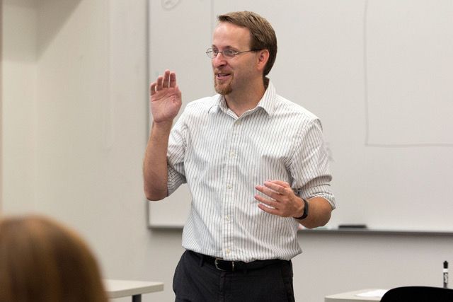 Male Professor lecturing in front of a class wearing a white button up and black slacks 