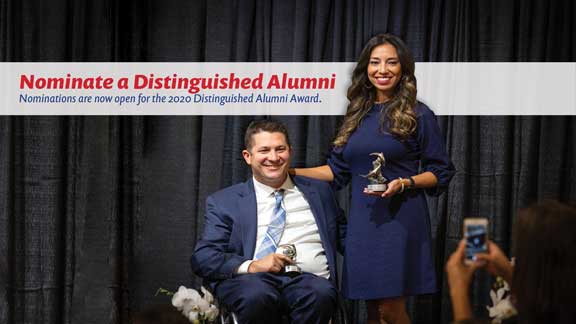 Man sitting down in a blue suit with a woman in a blue dress to his right holding an award