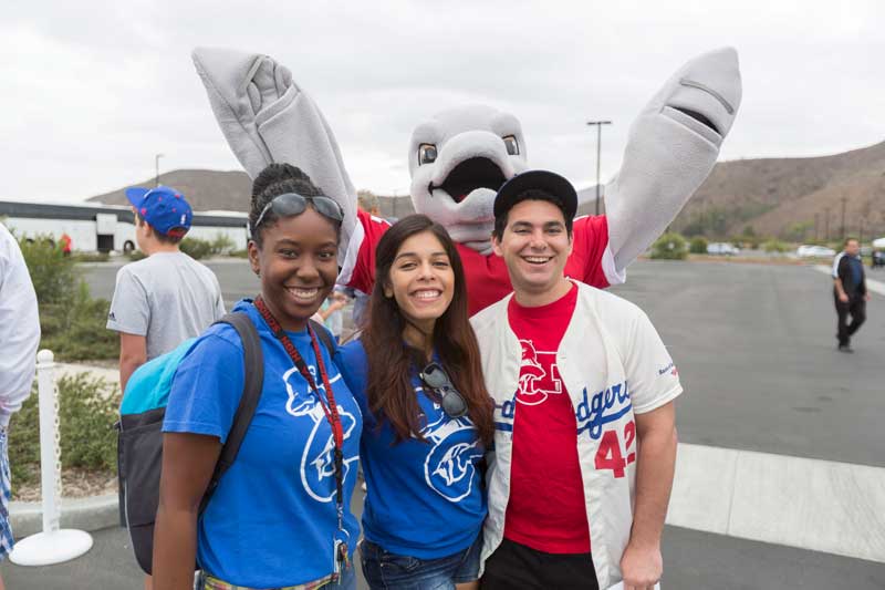 Three smiling CI Students wearing Dodger gear with mascot Ekho behind them