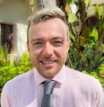 Smiling man in a pink shirt and gray tie, standing outdoors by tropical plants.