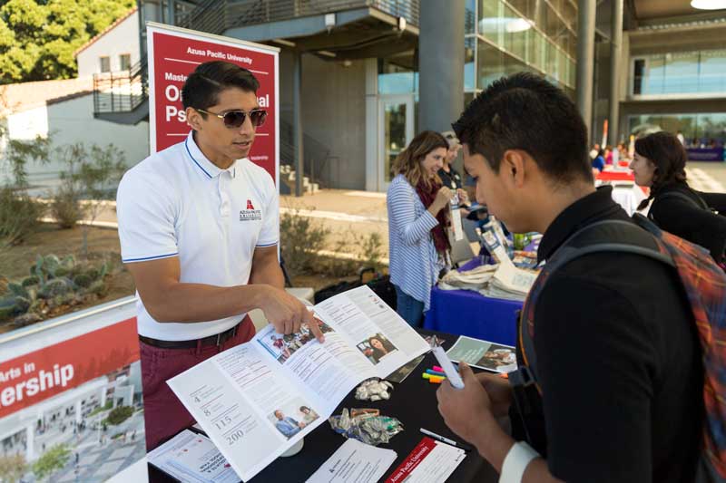 Two students at a career fair booth, student behind the booth in glasses, white polo, and burgundy pants is pointing and showing the student in front of the booth a pamphlet