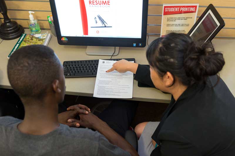 Two students sitting in front of a computer with a resume in front of them, student on the left is pointing to the resume