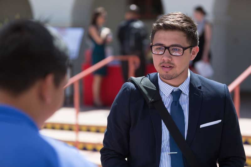 CI student wearing glasses in a suit and tie listening to a man wearing a blue shirt in front of him , with the Bell Tower building in the background