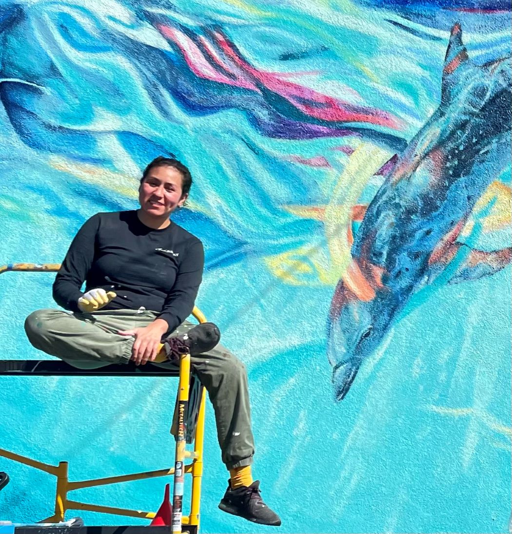 Muralist, Myisha Arellano muralist,Myisha Arellano, sitting on scafolding, backdrop camous legacy mural