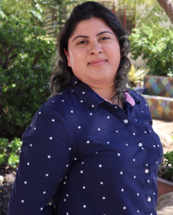 Christell Martinez, DAPI Secretary is smiling wearing a dark blue button up with polka dots. They have black hair. There re trees and other greenery in the background.