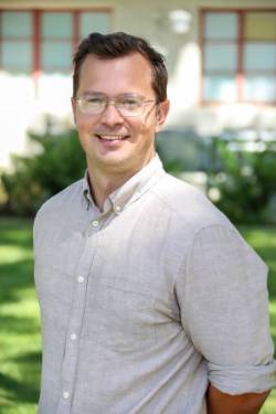 Dr. Kris Yeager, Faculty DAPI Advisor, is smiling with brown hair and wearing glasses. They are wearing a beige button up with the sleeves rolled up. There is greenery and a building with red trimmed windows in the background.