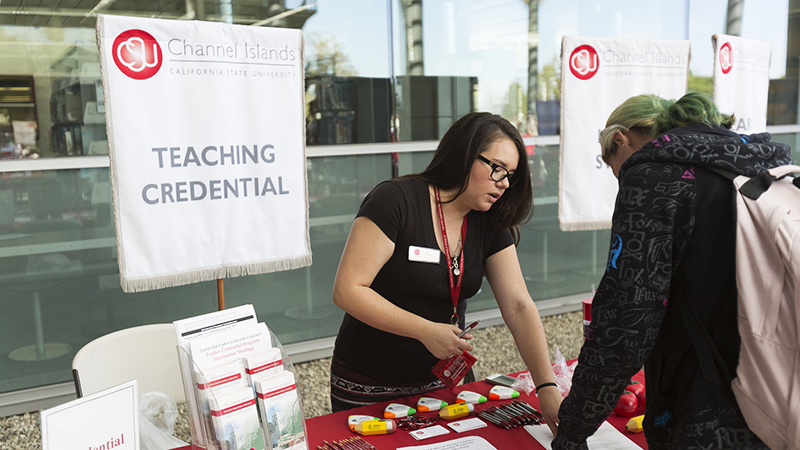 Teaching credential banner behind a booth with a woman showing a pamphlet to a person in front of the booth.