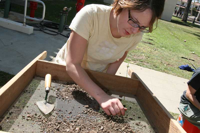 woman wearing glasses in yellow shirt using tools to inspect dirt sample