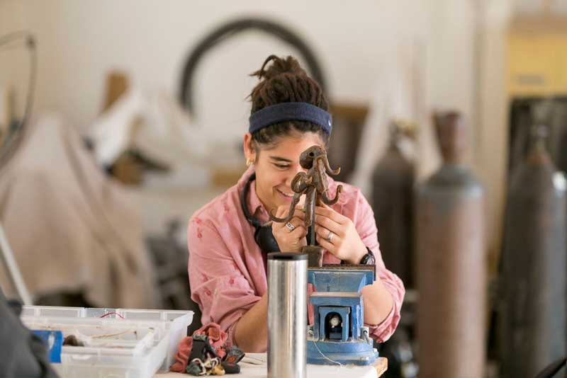 Female student in a blue headband and pink shirt working on a sculpture in an art classroom