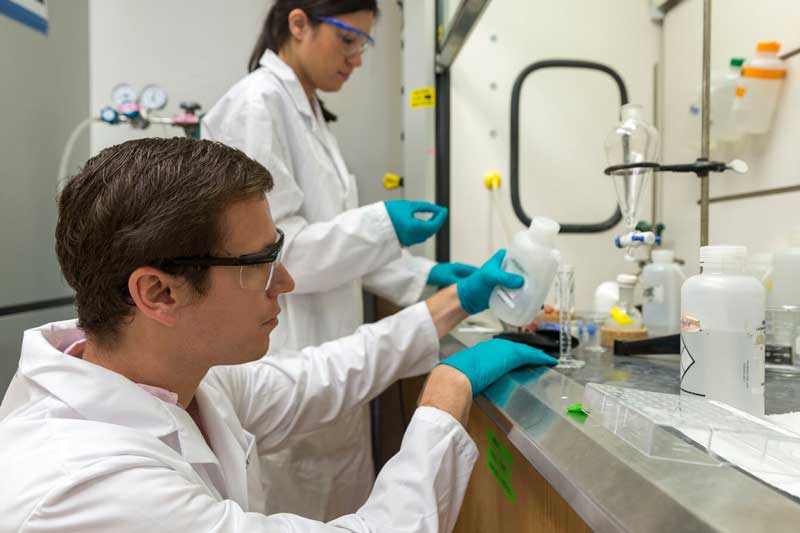A lab student in the background wearing a white lab coat, protective glasses and blue gloves inspecting a sample in her hand, a lab student in the foreground wearing a white lab coat, protective glasses and blue gloves pouring a solution into a glass test tube in a lab