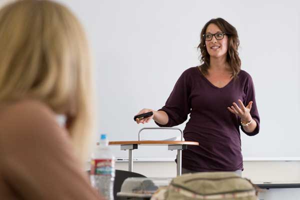 A lecturer wearing a maroon long sleeve in front of the class while a blonde is on the left corner focusing on the lecturer