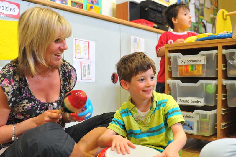 A blonde teacher in a classroom holding a rattle next to a young smiling boy wearing a lime green and blue striped polo.