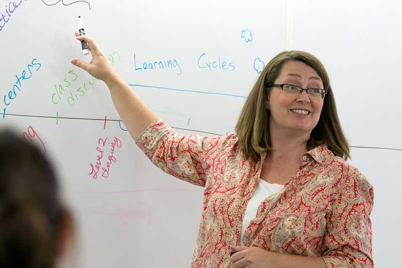 Lecturer wearing glasses smiling as she is pointing at a whiteboard with notes