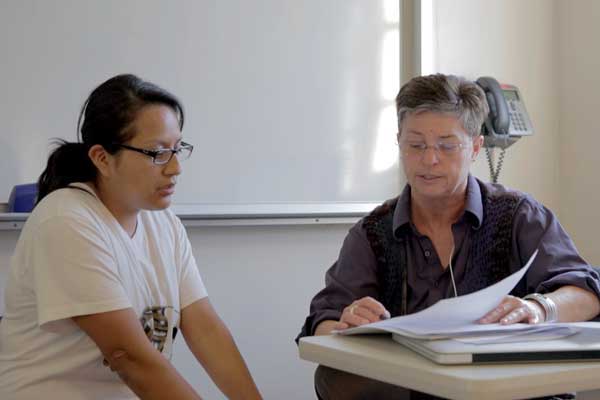 Woman wearing a white t-shirt looking at a paper together being proofread by a woman to her right