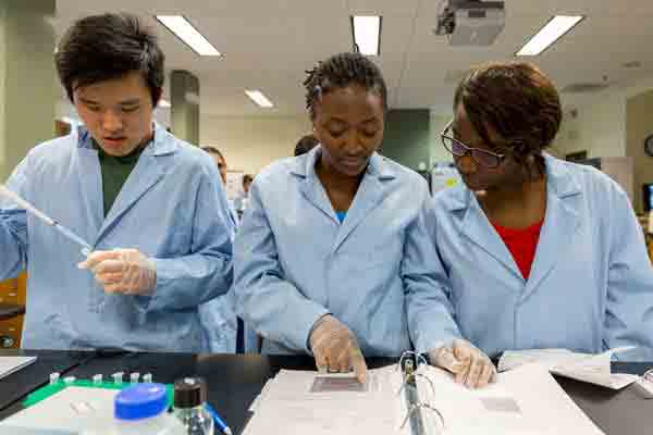 Three students in blue lab coats the one to the left pipng a sample and the two others reading a paper in a binder