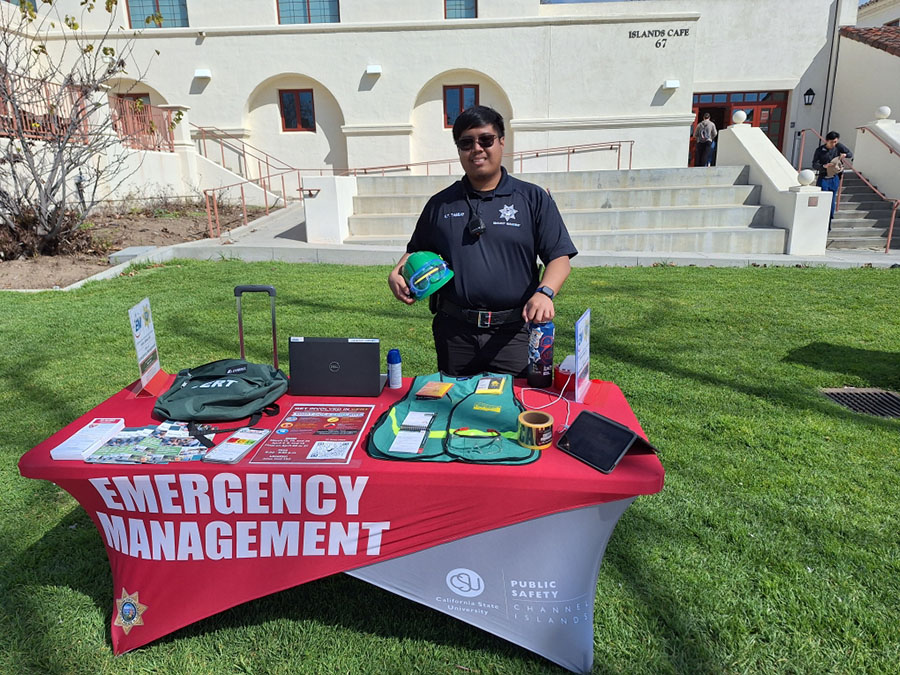 A young man stands at a table with emergency safety equipment and a red tablecloth that says "Emergency Management"