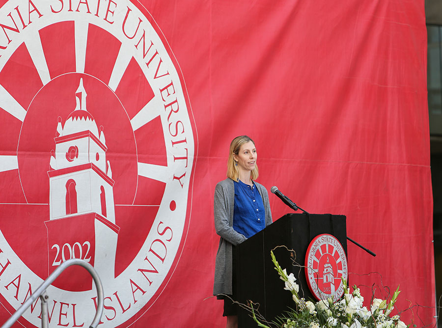 Melissa Soenke speaks at Convocation in front of a red backdrop with the CSUCI seal on it