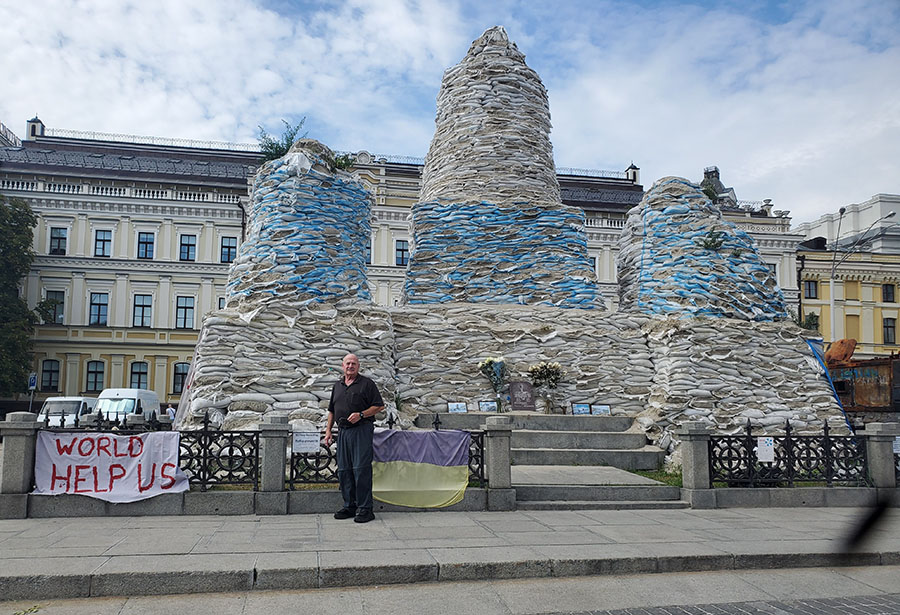 Dr. Baker stands in front of a 50-foot high stack of sandbags and a sign to the side that reads "World Help Us"