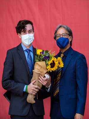 cameron and president yao stand together wearing masks in front of a red background. cameron is holding a bouquet of sunflowers and other flowers.