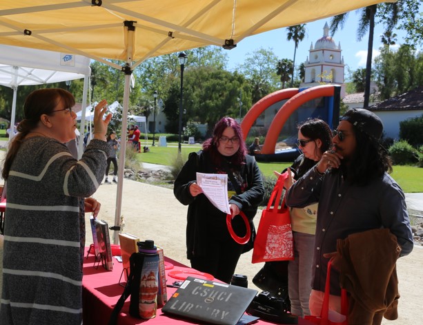Students are at the library resource table in front of the Bell Tower receiving information.