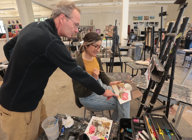 Student holding painting of a mug and donut, professor is standing near and pointing at painting