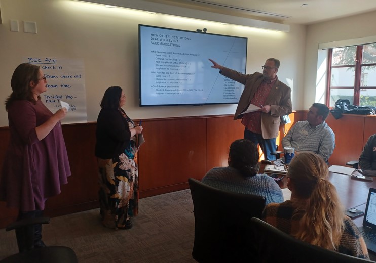 Three people presenting in front of a TV screen, one person is pointing at the screen.