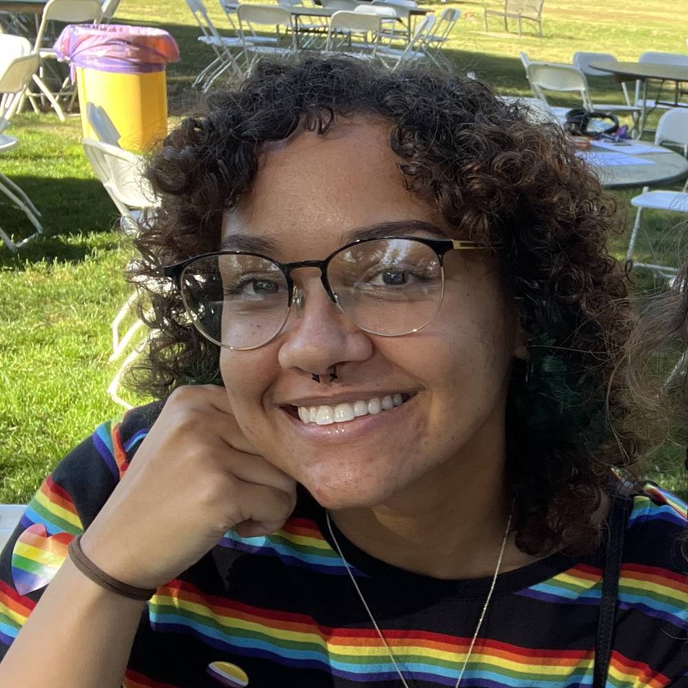 alva bailey has their chin rested on their hand. They are wearing square glasses and have short curly hair. 