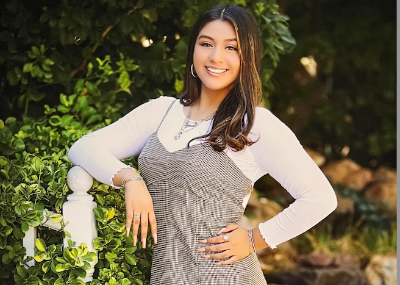 Isamar is standing and leaning against a white staircase. She is wearing a long white t-shirt and a checkered dress over it. She is smiling at the camera. 