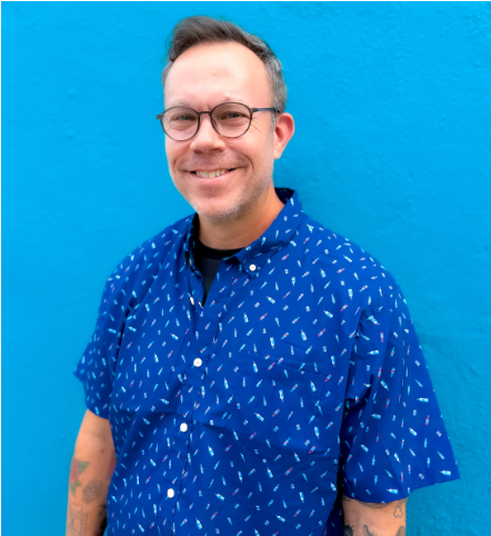 Jason is wearing black rimmed glasses, a blue button up, and is smiling directly at the camera. His background is a bright shade of blue. 
