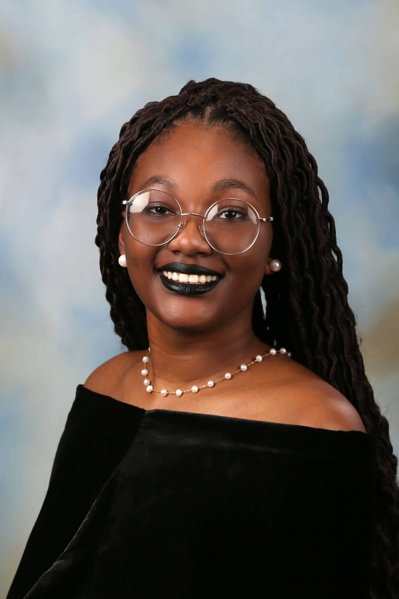 Jilllian newton has dark brown hair and has round silver glasses. She is wearing black lipstick with a black shoulder top and pearls. 
