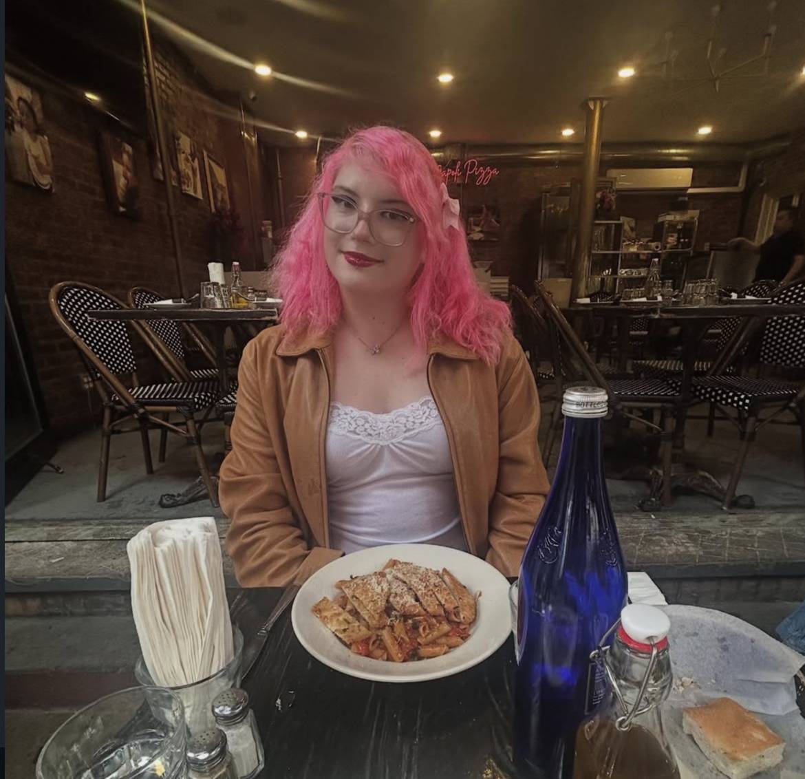 Scarlett has pink hair and is sitting at a restaurant eating pasta.