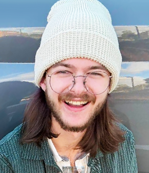 Zander is wearing a white knitted beanie, round glasses, and a green polo. He has long brown hair and a brown beard