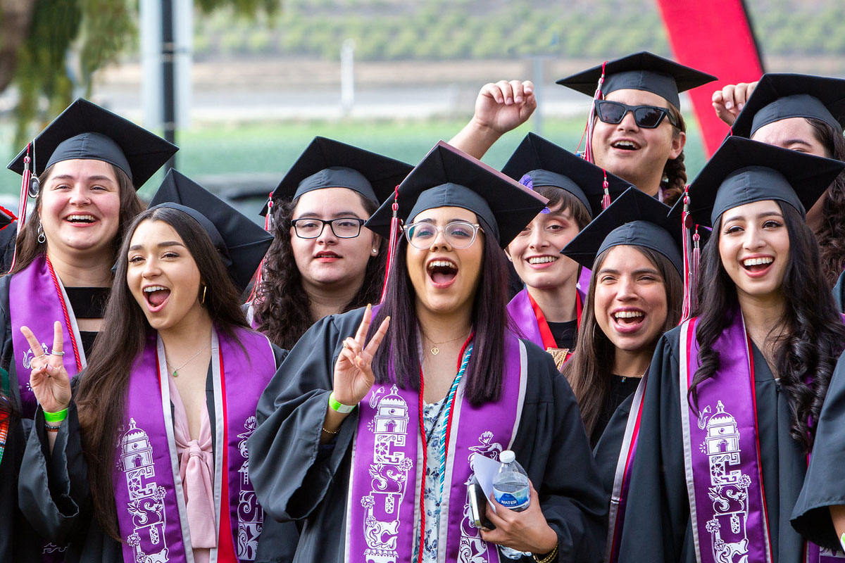 Group of smiling CSUCI graduates in black caps and gowns wearing purple stoles over a red stole
