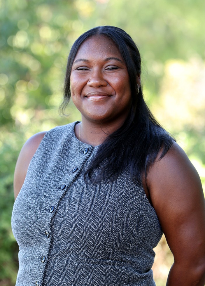 A smiling headshot of Loren wearing a gray vest against a blurred nature background.