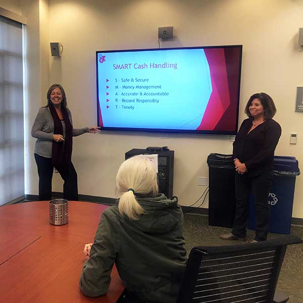 Meeting room with two presentors and a screen between then with a slide titled SMART Cash Handling and a woman sitting at a table in front of them watching the presentation