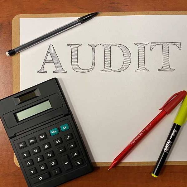 A white paper with the word Audit on a clipboard with a calculator to the bottom left and pens to the bottom right