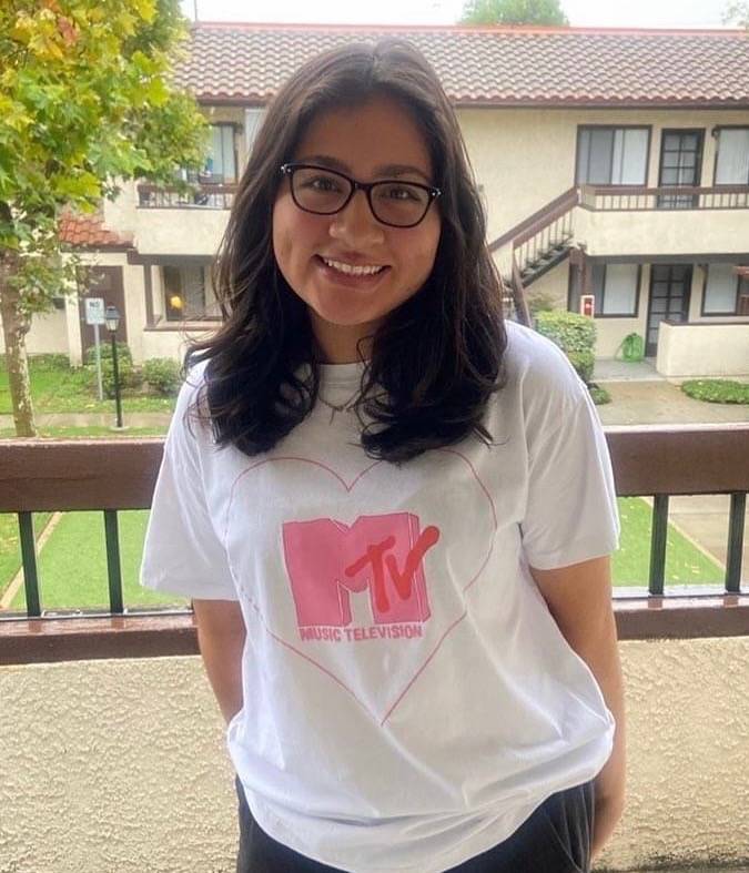 Alyssa has short dark hair, is wearing dark rimmed glasses, and is smiling at the camera. She is wearing a white shirt with pink lettering on it.