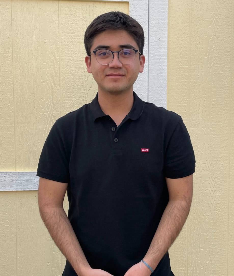 Carlos has short black hair. He is wearing black glasses and a black polo t-shirt. He is smiling at the camera and has his hands in front of him. 