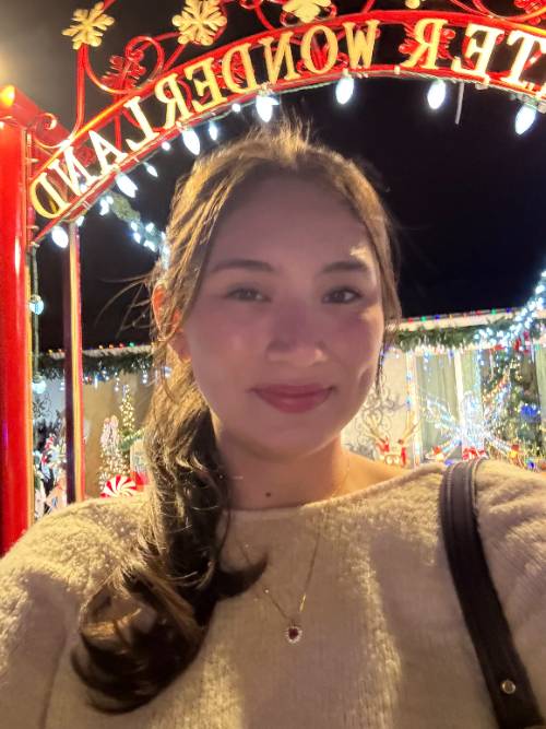 Trinity is smiling directly at the camera. She is wearing a light green sweater. She has long black hair and has a few strands in hair bands