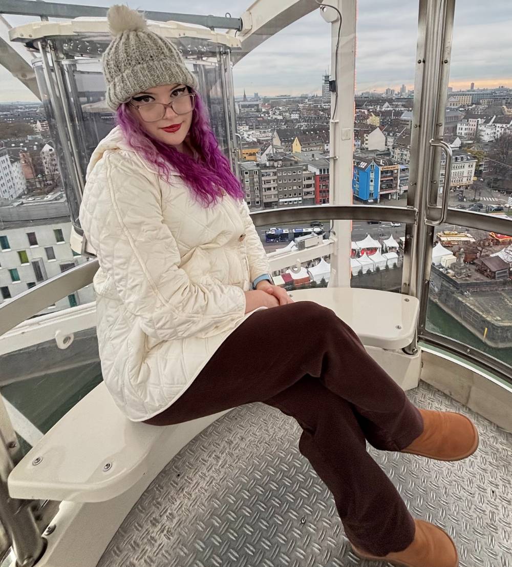Scarlett has bright red and curly hair with straight bangs. She is wearing a dark eyeliner wing and some red lipstick. She is smiling at the camera. 